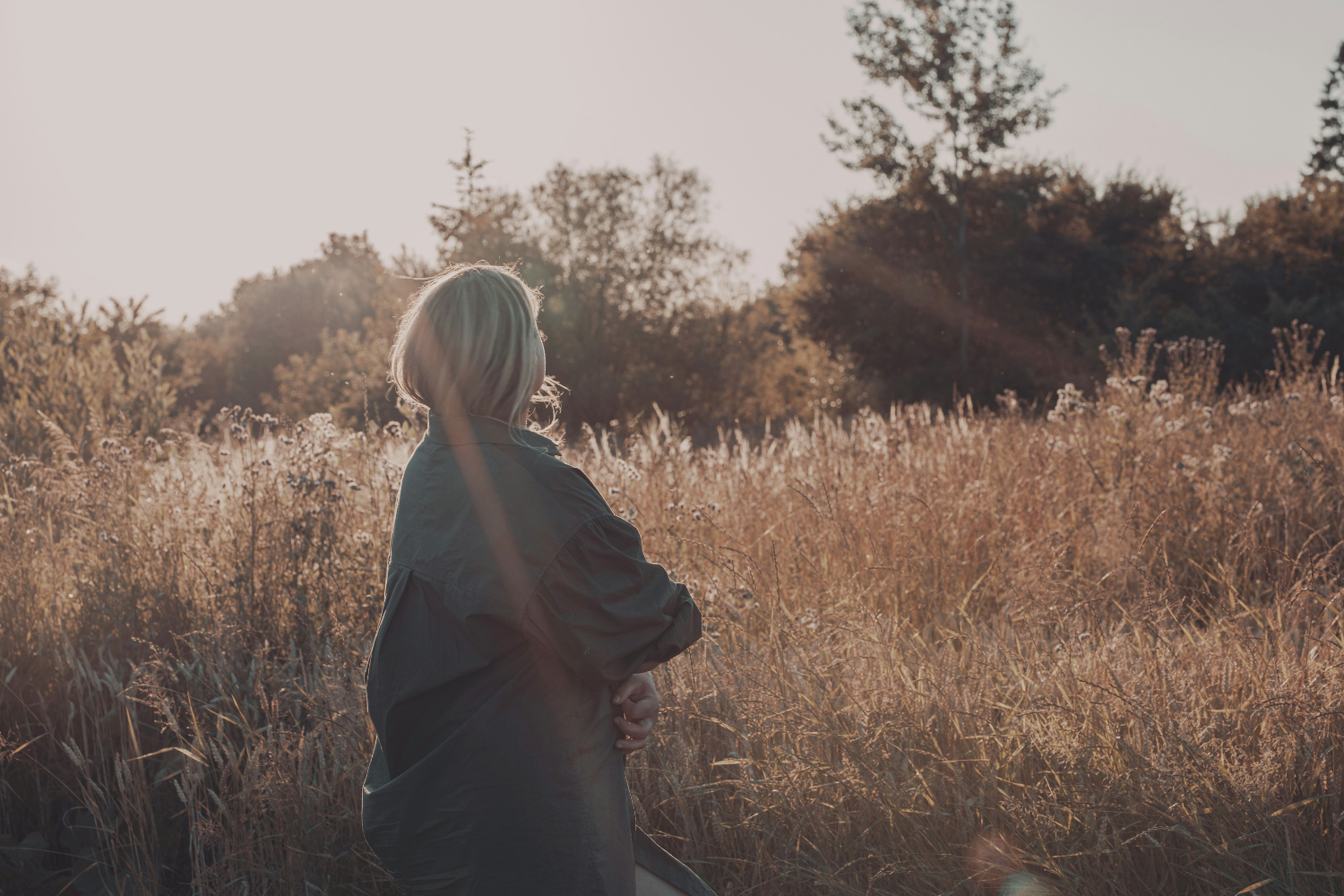 Vrouw van achteren gezien in een open veld met hoog gras, kijkend richting bomen in warm avondlicht.